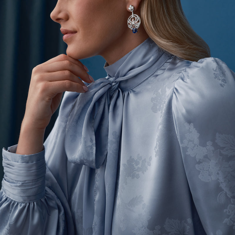 Woman wearing Royal Porcelain sapphire and diamond earrings in platinum, paired with a blue satin blouse.