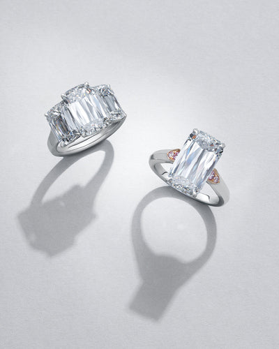 Two Ashoka diamond engagement rings with pink diamonds, modern classic trilogy design, on a light gray background