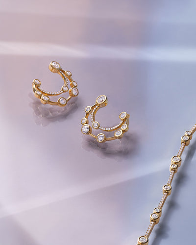 Gold crescent-shaped hoop earrings and a matching chain necklace, each adorned with small clear stones, arranged on a soft light gray surface.